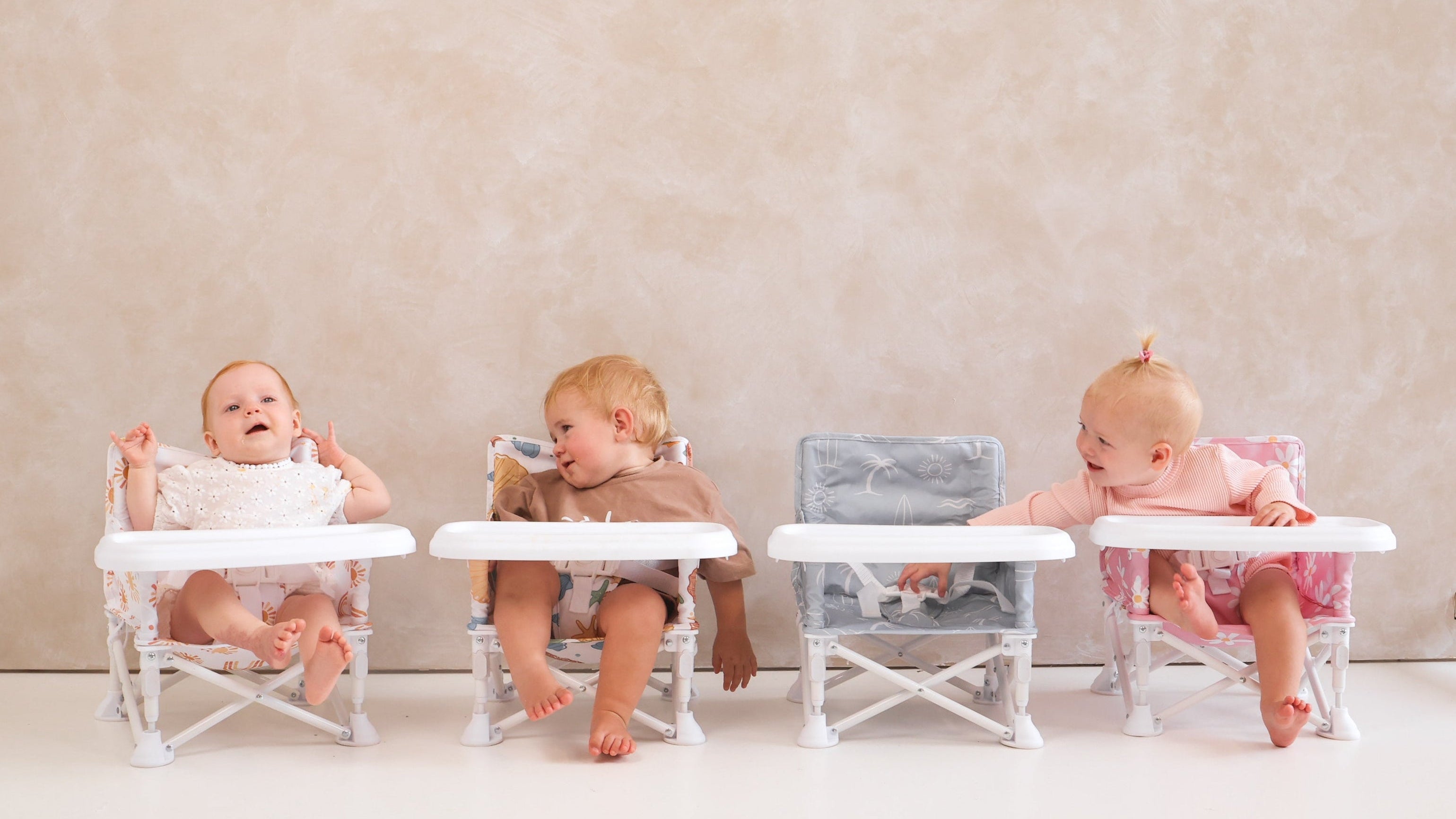 Three babies sitting in high chairs against a plain wall.