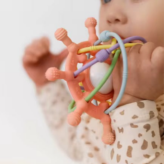 Baby chewing on the pink silicone teething toy, holding it with both hands.