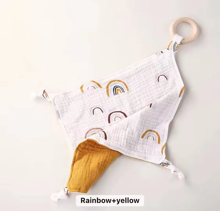 White muslin comforter with rainbow print and mustard backing, featuring a natural wooden teether ring.