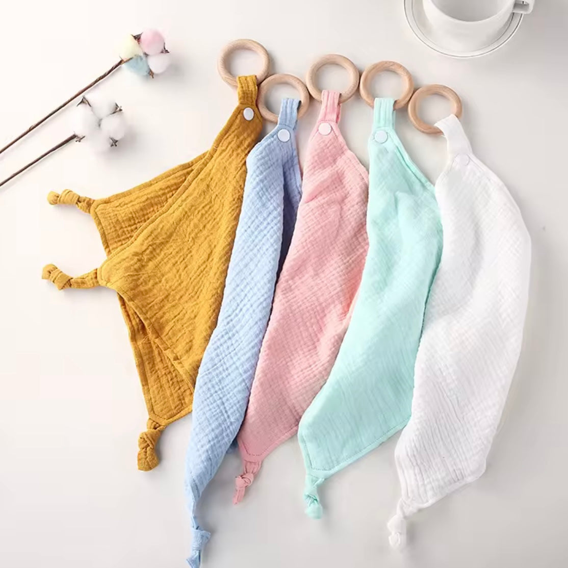 Set of five pastel muslin comforters with wooden teethers in mustard, baby blue, blush pink, mint, and white.