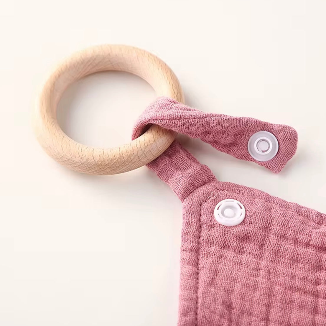 Close-up of natural wooden teething ring attached to soft dusty rose muslin comforter with secure snap closure.