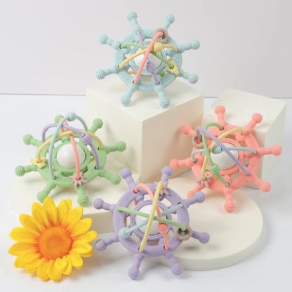 Group of four pastel baby teething toys in green, blue, purple, and pink, displayed on white blocks with a yellow flower for decoration.